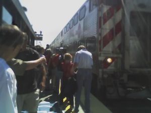 Riders boarding Caltrain in Mountain View station. Front of the train is on the right, with its lights on. Passengers boarding and alighting are on the left.
