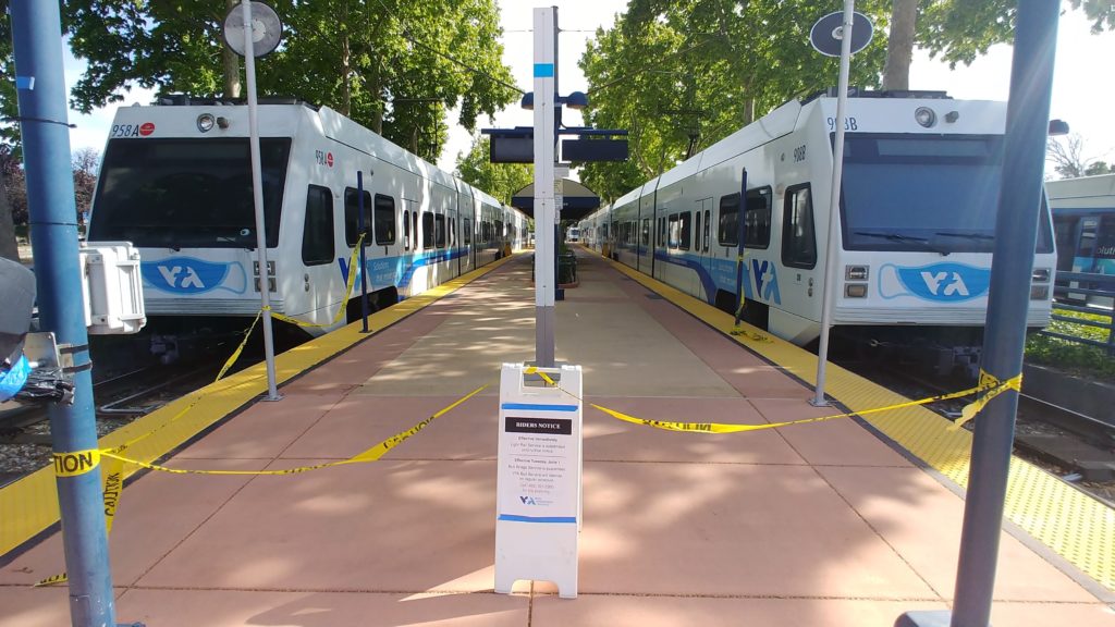 2 VTA light rail trains stopped at Santa Teresa station. Both trains are white, with the blue VTA logo with mask at the front. Camera is on the platfrom facing both stopped train cars.