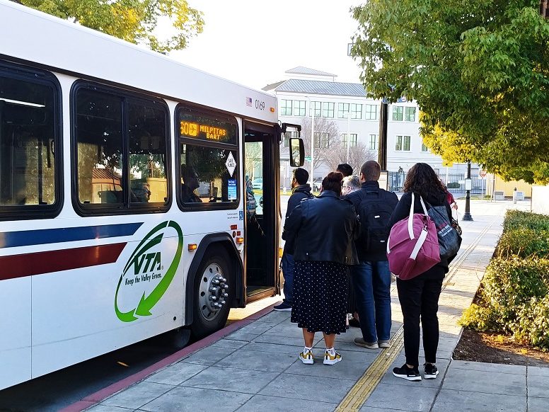 Passengers board VTA's 60 bus to San Jose Airport. Bus is white with burgundy and blue stripes. Six passengers are boarding the bus.