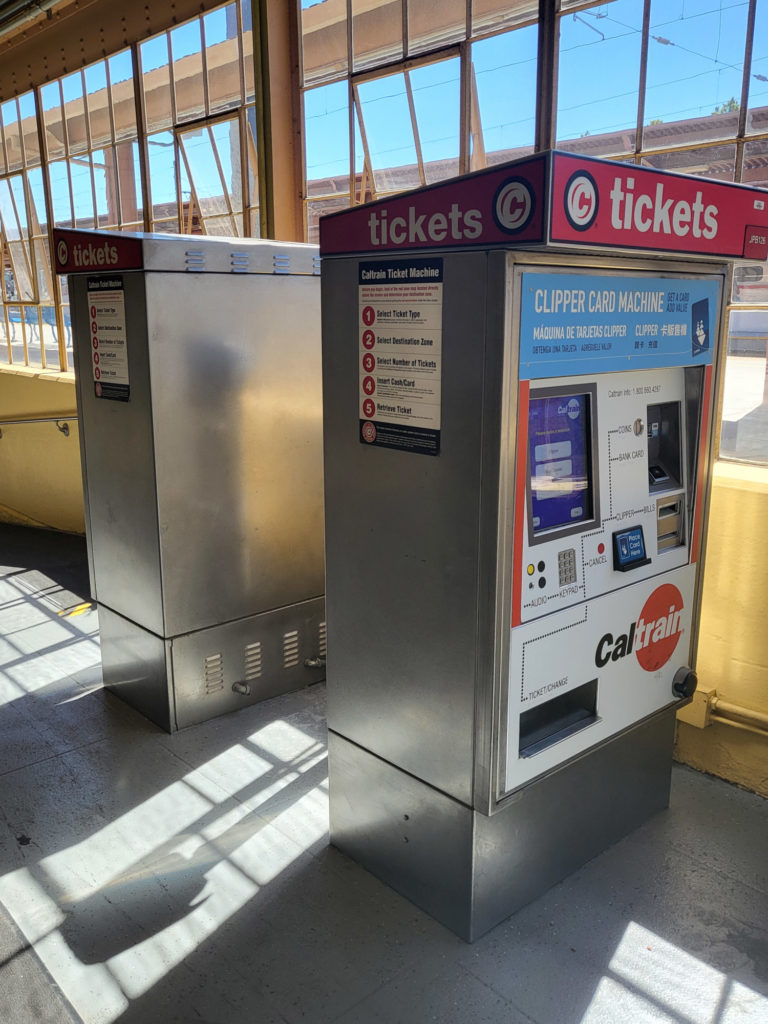 Two Caltrain ticket vending machines - back to back - inside San Jose Diridon Station. Caltrain TVMs have a red top with white customer-facing front. They have a blue strip over the screen that also says "Clipper Card Machine" where one can buy Clipper Cards to pay local or Bay Area regional transit fare.