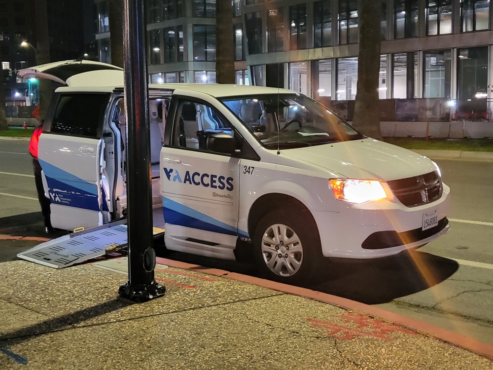 VTA ACCESS Paratransit van. A Dodge Caravan w/ wheelchair lift is parked on the street at night. Van is white with small blue stripes along the fender that flare towards the back. The van's lights are on.
